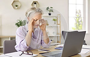 Unhappy gray haired woman suffers from headaches and migraines sits at table with laptop in office