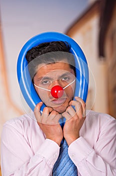 Unhappy handsome businessman with red plastic nose with a blue ballon aroun his head