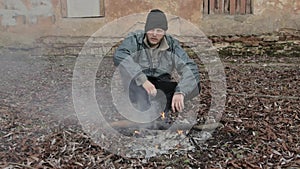 Unhappy homeless man sits by the fire. Sadness concept. Depressed tramp in dirty clothes and hat cap. Below poverty line