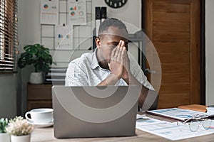 Unhappy hopeless African American man holding head in hands, overwhelmed tired businessman sitting at work desk with