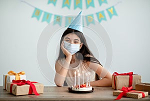 Unhappy Indian teenage girl in face mask and festive hat having dull birthday alone at home during covid isolation