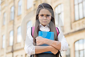 Unhappy little kid in eyeglasses hold study book in schoolyard outdoors, back to school