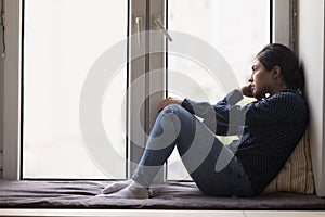 Unhappy lonely young Indian woman sitting on windowsill