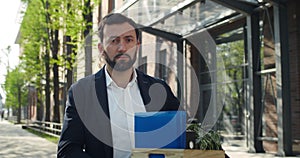 Unhappy man in 30s holding box with his belongings while standing at street. Male employee in formal suit turning head