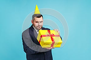 Unhappy man with beard wearing dark suit and party cone opening gift box and looking inside with