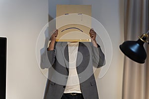 Unhappy man employee with box instead of his head. Portrait of businessman covering his face with paper mask with sad face drawn