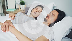 Unhappy man husband covering head with pillow while wife snoring in bed