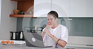 Unhappy man reading message with bad news on laptop computer, sitting at home kitchen. Shocked man scared with bad news