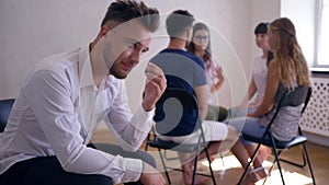 Unhappy man thinks about problems on group therapy session on background of people sitting on chairs in a circle