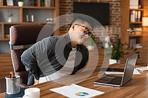 Unhappy middle aged asian man manager in glasses suffering from muscles back pain at table with laptop