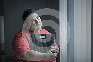 Unhappy obese man standing with scale by window