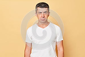 Unhappy offended man wearing white casual t-shirt standing isolated over beige background looking at camera with pout lips