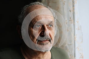 Unhappy old man indoors: senior man with grey beard and moustache standing near window. Loneliness, aged people problems concept