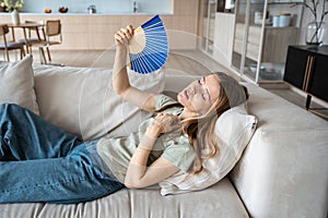 Unhappy overheated woman lying on sofa at home waving paper fan to cool down suffers from stuffiness