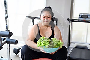 Unhappy overweight Asian female looking for organic green vegetable salad in gym. Asian women in sportswear do not want to eat