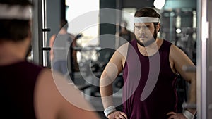 Unhappy overweight man looking at his mirror reflection in gym, diet and sport