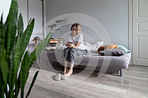 Unhappy pessimistic woman sits on bed with phone in hands waiting for call from boyfriend or husband