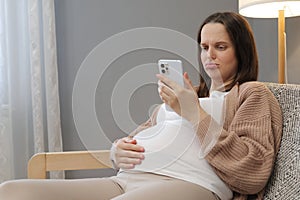 Unhappy pregnant woman using smartphone at home browsing pregnancy app while sitting on couch in living room smiling expectant