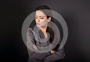 Unhappy resentful business woman in suit with folded arms looking down on dark grey background