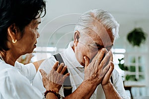 Unhappy retired Asian senior couple sitting on the sofa while elderly wife comforting her sad husband at home.