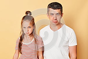 Unhappy sad upset man and little kid girl standing together isolated over beige background looking at camera with pout lips being