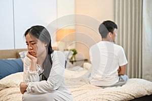 An unhappy, sad young Asian couple in pajamas is sitting separately on the bed after their argument