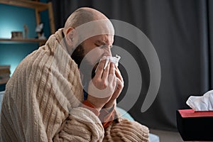 Unhappy sad young man in plaid suffering from fever and flu on sofa, blowing nose in napkin. Treatment of illness, cold