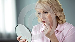 Unhappy senior female looking at sagging skin face in mirror, old age appearance