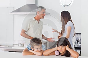 Unhappy siblings sitting in kitchen with their parents who are f