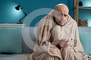 Unhappy sick man sick at home sitting with thermometer on sofa. Virus and high fever concept