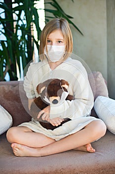 Unhappy sick toddler girl wearing medicine mask for protection sitting with toy teddy bear