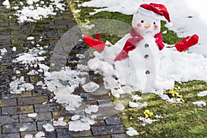 Unhappy snowman in mittens, red scarf and cap is melting  outdoors in sunlight on snowy green grass with small yellow flowers near