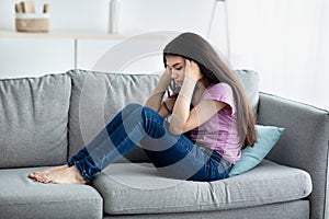 Unhappy teen girl holding her head in despair, feeling depressed, sitting on sofa at home, copy space