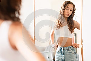 Unhappy teen girl looking critically at herself in mirror indoors