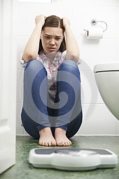 Unhappy Teenage Girl Sitting On Floor Looking At Bathroom Scales