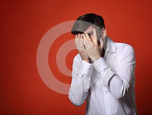 Unhappy thinking grimacing bearded business man covering the face his hands in white shirt and looking on bright orange