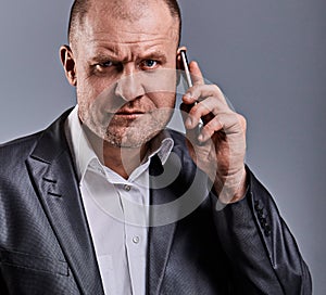Unhappy tired angry business man talking on mobile phone and holding in hand one more phone in office suit on grey studio