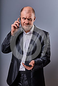 Unhappy tired angry business man talking on mobile phone and holding in hand one more phone in office suit on grey studio