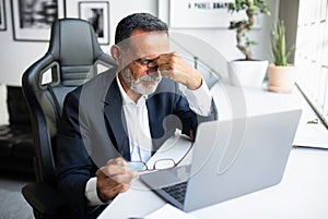 Unhappy tired european senior businessman in suit resting at table with computer in office