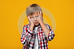 Unhappy upset disgruntled caucasian teenage boy pupil presses hands to face, isolated on yellow background