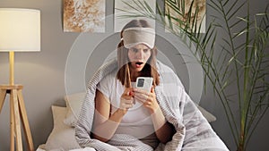 Unhappy woman in blindfold holding modern smart phone chatting online reading bad shocked news poses on bed wrapped in striped