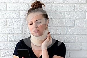 Unhappy woman with cervical surgical collar and smartphone feeling pain in neck against a blurred white brick wall