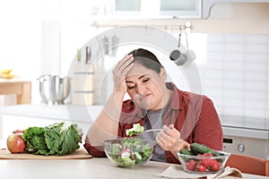 Unhappy woman eating vegetable salad at table in kitchen