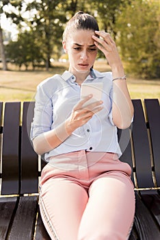 Unhappy woman sitting on bench in autumn park with mobile phone