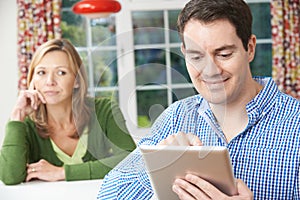 Unhappy Woman Sitting At Table As Partner Uses Digital Tablet