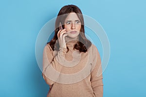 Unhappy worried woman talking on phone isolated over blue background, wearing casual beige turtleneck, looks away with scared