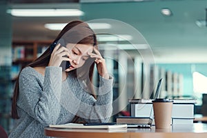 Unhappy young asian woman talking on mobile phone while reading book in library