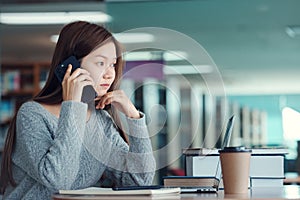 Unhappy young asian woman talking on mobile phone while reading book in library