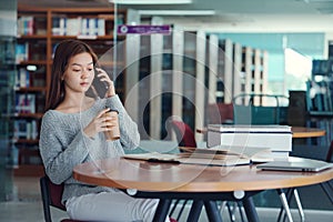 Unhappy young asian woman talking on mobile phone while reading book in library