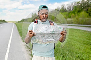 Unhappy young black man hitchhiking on road, looking at map, feeling lost, traveling alone by autostop